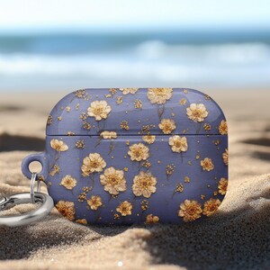 May include: A blue case for wireless earbuds, adorned with a pattern of dried yellow flowers. The case has a silver carabiner clip attached. The background shows a sandy beach and the ocean.