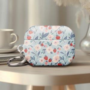 May include: A light blue AirPod case with a floral pattern of strawberries and pink flowers. The case has a silver carabiner clip attached. A white teacup and a book are in the background.