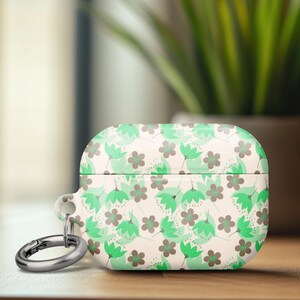 May include: A white and green floral patterned AirPod case with a silver carabiner clip. The case features a repeating pattern of green leaves and brown flowers. The case is on a wooden surface.