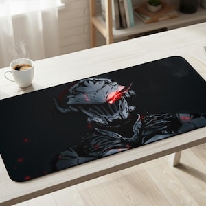 May include: Black rectangular desk mat with a detailed illustration of a character with a helmet and glowing red eyes. The design is set against a dark background. A coffee cup and a small plant are on the table.