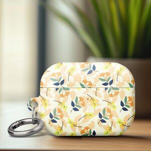 May include: A white AirPods case with a floral pattern featuring orange, yellow, and blue leaves and flowers. The case has a silver carabiner clip attached. The case is on a wooden surface.