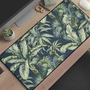 May include: A rectangular desk mat featuring a tropical foliage design. The mat showcases a dark blue background with detailed illustrations of green and light green palm leaves and other tropical plants. The mat is rectangular and has a black border.