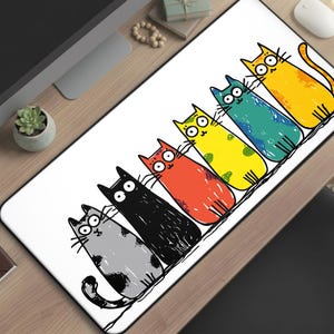 May include: A white rectangular desk mat featuring a cartoon illustration of six colorful cats. The cats are in various colors including gray, black, red, yellow, blue, and orange. The design is outlined in black.