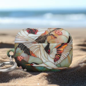 May include: AirPods case with a crane design. The case features a colorful illustration of a crane in flight, with white, orange, and black details. It has a silver carabiner clip attached.