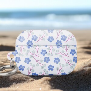 May include: White AirPods case with a floral pattern of blue flowers and pink stems. The case has a silver carabiner clip attached. The background is a sandy beach with the ocean in the distance.