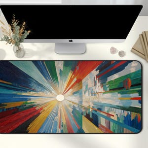 May include: A rectangular desk mat with a vibrant abstract design. The artwork features a central white circle with radiating lines in shades of red, blue, green, and yellow. The mat is on a white desk with a computer monitor and other desk accessories.