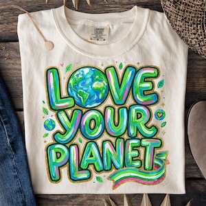 May include: Cream-colored t-shirt with the words "LOVE YOUR PLANET" in green, blue, and pink. The design includes a globe and decorative leaves. The shirt is laid out with blue jeans, sunglasses, a straw hat, and boots on a wooden surface.