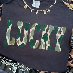 May include: Black t-shirt with the word "LUCKY" in a camouflage pattern. A gold necklace with charms and sunglasses are on top of the shirt. The shirt is on a leopard print surface.