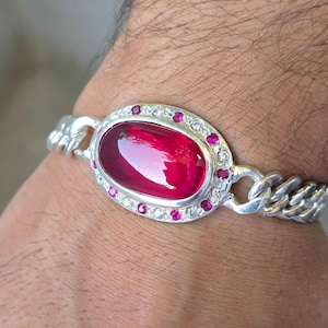 Może przedstawiać: Srebrna bransoletka łańcuszkowa z dużym, owalnym, ciemnoczerwonym kamieniem szlachetnym typu kaboszon pośrodku. Centralny kamień otoczony jest aureolą małych, naprzemiennie jasnobiałych i jaskraworóżowych okrągłych kamieni w srebrnej oprawie.