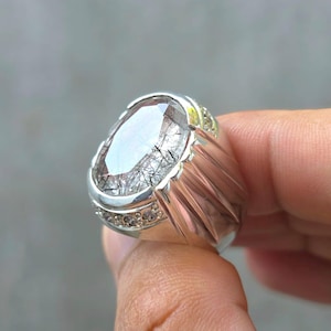 Pode incluir: Um anel de prata com uma grande pedra preciosa oval. A pedra preciosa transparente tem inclusões pretas, e o anel tem um design canelado nas laterais e pequenas pedras ao redor da pedra principal.