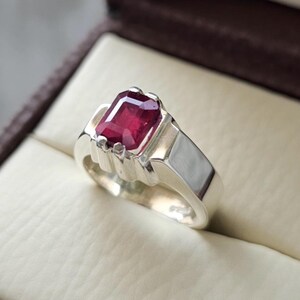 Puede incluir: Un anillo de plata con una piedra preciosa de color rojo intenso, tallada en forma rectangular y engastada en una montura de garras. El aro es liso y pulido, con una estructura de metal ancha y sólida que se estrecha ligeramente.