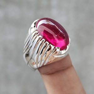 May include: A silver ring featuring a large, oval-shaped, deep pinkish-red cabochon gemstone. The metal band displays a series of vertical, grooved, and textured ridges that extend from the central stone setting down the sides of the ring.