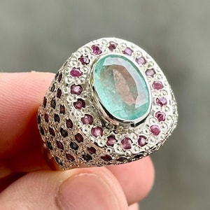 May include: A silver ring featuring a large, oval-cut, light green gemstone at the center, surrounded by smaller, round, dark pink gemstones. The ring has a textured, detailed band.