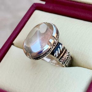 Pode incluir: Um anel de prata com uma pedra preciosa oval e transparente. O anel tem uma banda de prata detalhada com padrões gravados e um sotaque escuro. O anel é apresentado numa caixa de joias bordô com um interior creme.