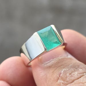 May include: A silver ring with a rectangular, emerald-cut, bright green gemstone. The ring has a wide band and a polished finish, showcasing the vibrant color of the emerald. The setting appears to be a combination of silver and gold.