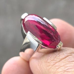 Op de afbeelding: Een zilveren ring met een grote, ovaalvormige, dieprode edelsteen. De edelsteen is het middelpunt, gezet in een gepolijste zilveren band met een modern, geometrisch ontwerp. De zetting van de ring heeft een minimalistische stijl.