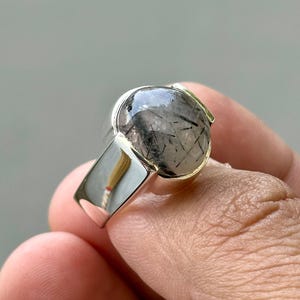 Pode incluir: Um anel de prata com uma grande pedra redonda com inclusões pretas. A pedra preciosa é engastada em uma banda de prata polida. O anel é segurado por uma mão, mostrando o design e os detalhes.