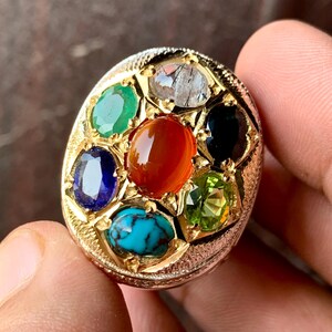 May include: A gold ring featuring a central oval orange carnelian stone surrounded by six smaller faceted gemstones including a green emerald, a blue sapphire, a turquoise, a clear rutilated quartz, a black onyx, and a light green peridot.