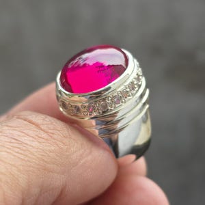 May include: A silver ring featuring a large, oval-shaped, vibrant red gemstone. The band is accented with a row of smaller, clear stones. The ring has a polished, reflective surface, and the gemstone has a glossy appearance.