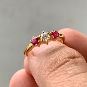 May include: A gold ring featuring a central clear round-cut gemstone flanked by two smaller round-cut red rubies. The stones are set in a prong setting on a polished gold band, creating a three-stone design with a classic and elegant appearance.
