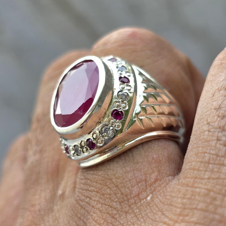 May include: Silver ring featuring a large oval ruby gemstone. The ring is accented with smaller red gemstones and clear stones, set in a detailed silver band. The design includes engraved patterns on the sides, creating a classic and elegant look.