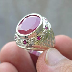 May include: A silver ring featuring a large, oval-cut ruby gemstone as the centerpiece. The ring is accented with smaller rubies and clear gemstones around the band. The band has a textured, geometric design.