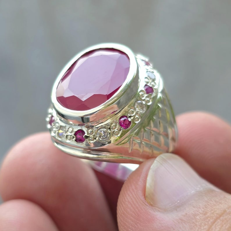 May include: A silver ring featuring a large, oval-cut ruby gemstone as the centerpiece. The ring is accented with smaller rubies and clear gemstones around the band. The band has a textured, geometric design.