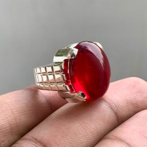 Può includere: Un anello color argento con una grande pietra preziosa traslucida rosso scuro di forma ovale, incastonata in una montatura in metallo lucido. La fascia dell'anello presenta un motivo a griglia testurizzato sul lato, dall'aspetto strutturato.