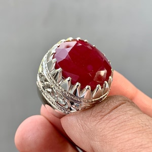 Peut inclure: Gros anneau avec une pierre précieuse rouge rubis ovale. La pierre est taillée pour refléter la lumière. La bague a un anneau argenté avec des détails complexes autour de la pierre.