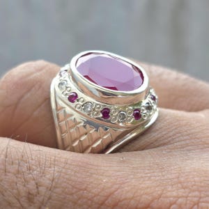 May include: A silver ring featuring a large, oval-cut ruby gemstone. The ring is accented with smaller red and clear gemstones around the band. The band has a textured, geometric design.