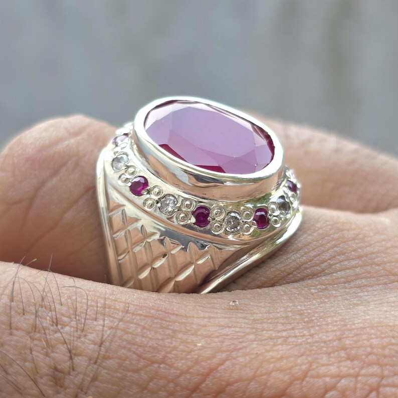 May include: A silver ring featuring a large, oval-cut ruby gemstone. The ring is accented with smaller red and clear gemstones around the band. The band has a textured, geometric design.