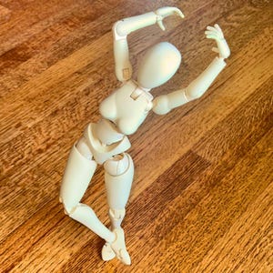 Op de afbeelding: Een crèmekleurig, gearticuleerd houten kunstenaarsmannequin in een dynamische pose, met opgeheven armen en gebogen benen. Het mannequin staat op een houten oppervlak en toont zijn gelede structuur voor poseren en schetsen.