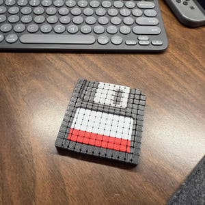 Puede incluir: Un posavasos de disquete impreso en 3D con un diseño pixelado. El posavasos presenta un borde gris, un centro blanco y rojo y un cuadrado gris en la esquina superior derecha. Se encuentra sobre una superficie de madera cerca de un teclado.