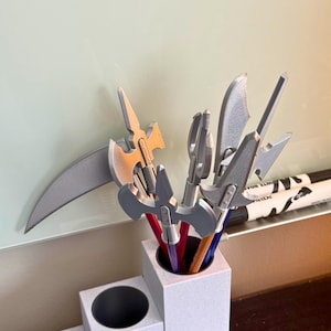 May include: A white desk organizer with a cylindrical pencil holder and a smaller compartment. The pencil holder contains pencils topped with miniature weapon-shaped toppers in silver and gold. A whiteboard and markers are in the background.