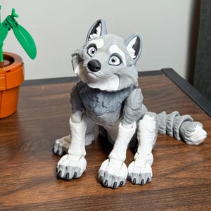Puede incluir: Figura de lobo gris y blanco con una expresión amigable. El lobo tiene ojos grandes y expresivos y una textura de pelaje detallada. Está sentado sobre sus cuartos traseros, con la cola enroscada. La figura está hecha de un material sólido y es un objeto decorativo.
