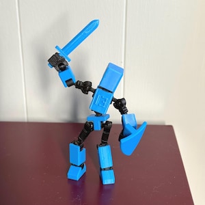 Puede incluir: Una figura de robot de juguete articulada azul con una espada y un escudo. El robot tiene un diseño de bloques y está en posición de combate.