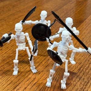 Könnte beinhalten: Fünf weiße Skelettfiguren aus Kunststoff mit schwarzen Waffen und Schilden. Die Skelette sind in Kampfstellung.