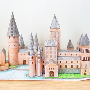 Peut inclure: Maquette en papier détaillée d'un château avec de multiples tours et flèches. Le modèle présente un schéma de couleurs beige, gris et marron, avec un pont et des accents verts. Le château est posé sur une base bleue et verte.