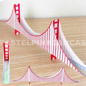 May include: A paper model of the Golden Gate Bridge, featuring a red and white color scheme. The bridge design includes vertical red lines and a road with yellow lines. The model is displayed on a light brown surface.