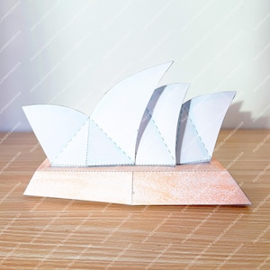 May include: A paper model of the Sydney Opera House, with white sail-shaped roofs on a tan base. The model is made from folded paper, representing the famous architectural design. The image is set on a wooden surface.