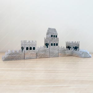 Può includere: Un modello di carta grigia di una sezione della Grande Muraglia cinese. Il modello presenta una torre centrale e due torri più piccole, con mura merlate e finestre ad arco. Il modello è realizzato in carta grigia e poggia su una superficie marrone chiaro.