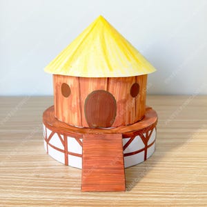 Könnte beinhalten: Ein Papiermodell eines kleinen Hauses mit gelbem Kegeldach, braunen Wänden und einer braunen Tür. Das Haus steht auf einem weiß-braunen Sockel mit einer Holzrampe, die hinaufführt. Das Design erinnert an eine Illustration aus einem Bilderbuch.
