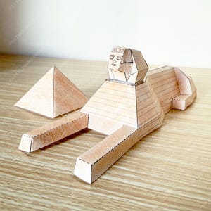 May include: A paper craft Egyptian Sphinx and pyramid set. The Sphinx is tan with a detailed face and body, and the pyramid is a simple triangular shape. The set is designed to be assembled, showcasing a miniature ancient Egyptian theme.