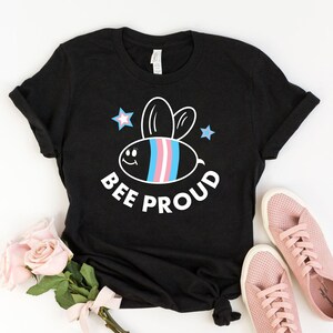 以下が含まれることがあります： 白い漫画の蜂が描かれた黒いTシャツ。蜂の縞模様にはトランスジェンダーのプライドカラーが使われています。蜂の周りは白い星で囲まれており、蜂の下には「BEE PROUD」という文字があります。