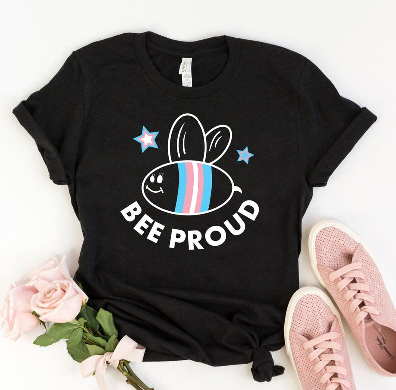 以下が含まれることがあります： 白い漫画の蜂が描かれた黒いTシャツ。蜂の縞模様にはトランスジェンダーのプライドカラーが使われています。蜂の周りは白い星で囲まれており、蜂の下には「BEE PROUD」という文字があります。