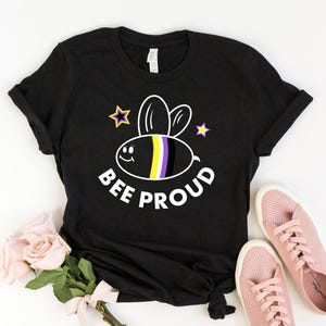 Könnte beinhalten: Schwarzes T-Shirt mit einer Cartoon-Biene mit gelben, weißen und violetten Streifen. Die Biene hat ein lächelndes Gesicht und ist von Sternen umgeben. Der Text "BEE PROUD" ist unter der Biene gedruckt.