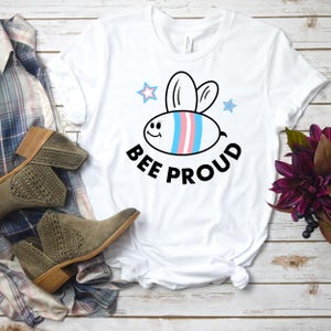 以下が含まれることがあります： 白いTシャツに、漫画の蜂のデザインがプリントされています。蜂は青、ピンク、白のストライプで、笑顔をしています。蜂の下には「BEE PROUD」という文字がプリントされています。