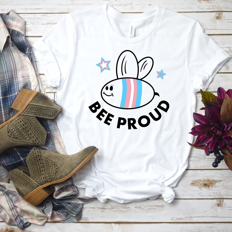 以下が含まれることがあります： 白いTシャツに、漫画の蜂のデザインがプリントされています。蜂は青、ピンク、白のストライプで、笑顔をしています。蜂の下には「BEE PROUD」という文字がプリントされています。
