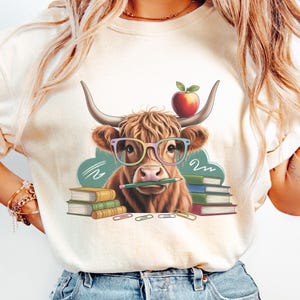 May include: A cream-coloured t-shirt with a cartoon illustration of a Highland cow wearing glasses and holding a pencil. An apple sits on its head, with books and paperclips completing the school theme. The cow is brown with long horns.