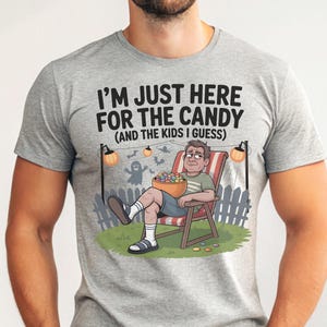 May include: Gray t-shirt with the text "I'M JUST HERE FOR THE CANDY (AND THE KIDS I GUESS)". The graphic shows a cartoon person relaxing in a chair, holding a bowl of candy, with Halloween decorations.